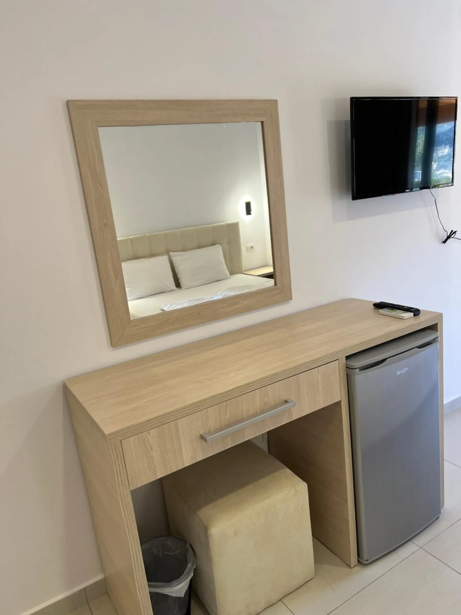 An oak desk and mirror with a small fridge and a stool, and a television on the wall