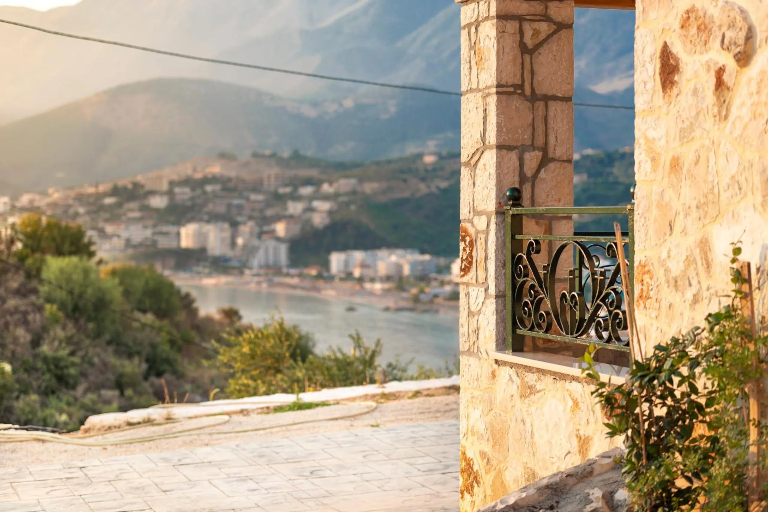 Late sun on a stone pillar and an iron balcony rail, with the bay and the town of Himarë soft in the haze beyond