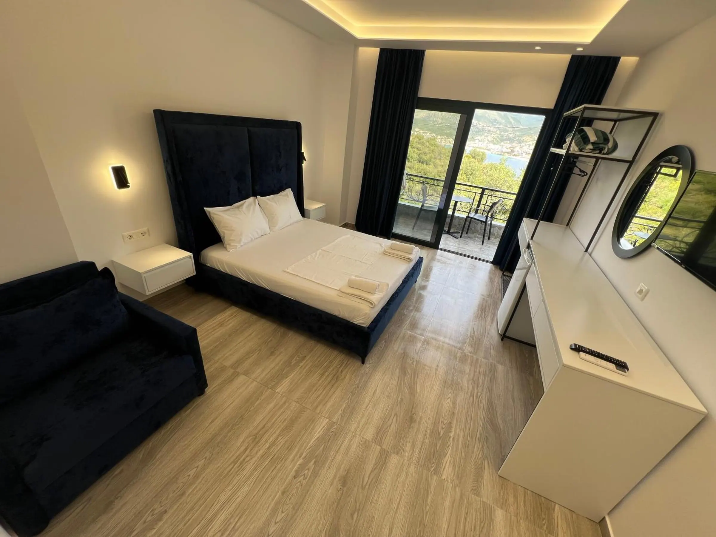 A double with a tall dark velvet headboard, a sofa, a white desk and a balcony door at the far end