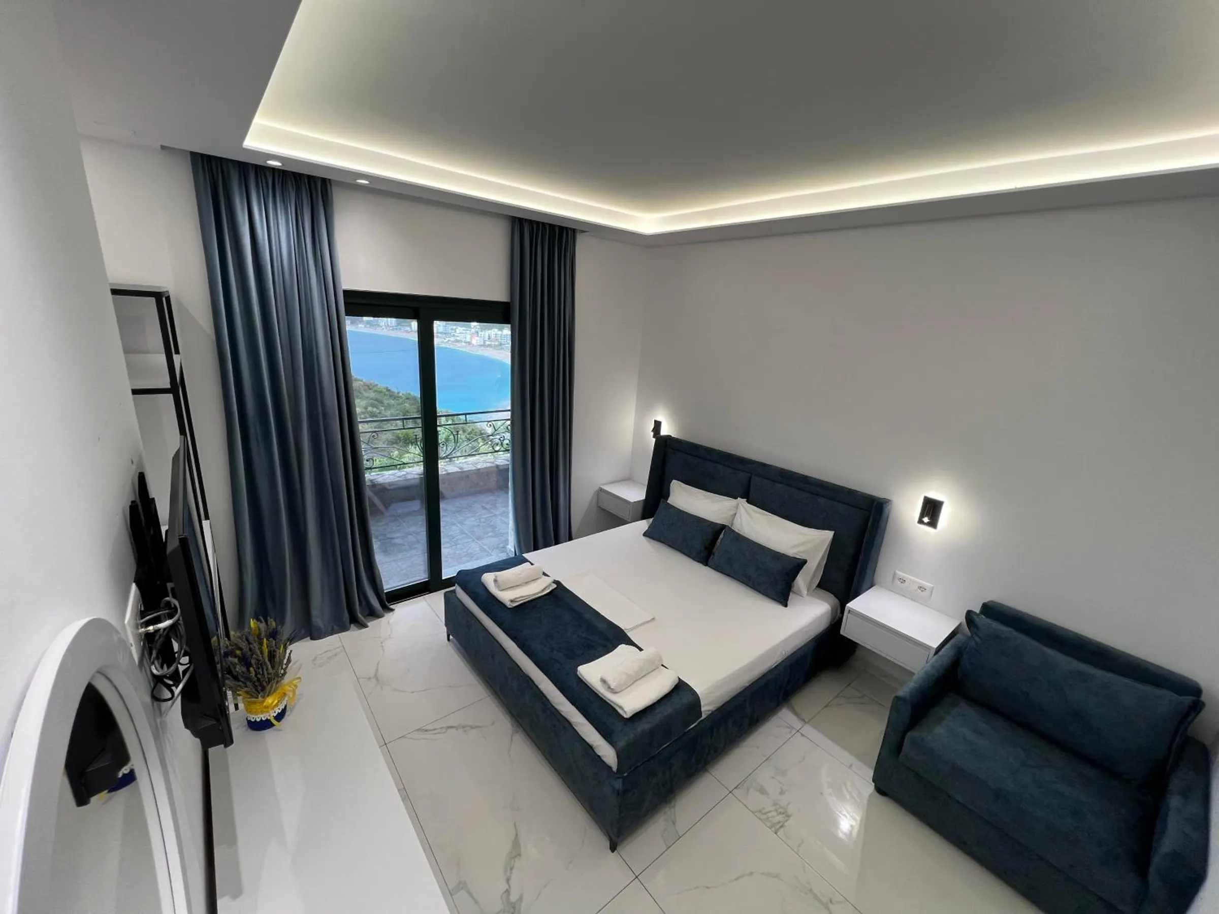 A double bed in navy velvet and a velvet armchair on a white marble floor, with a tall glass door onto the balcony and the bay