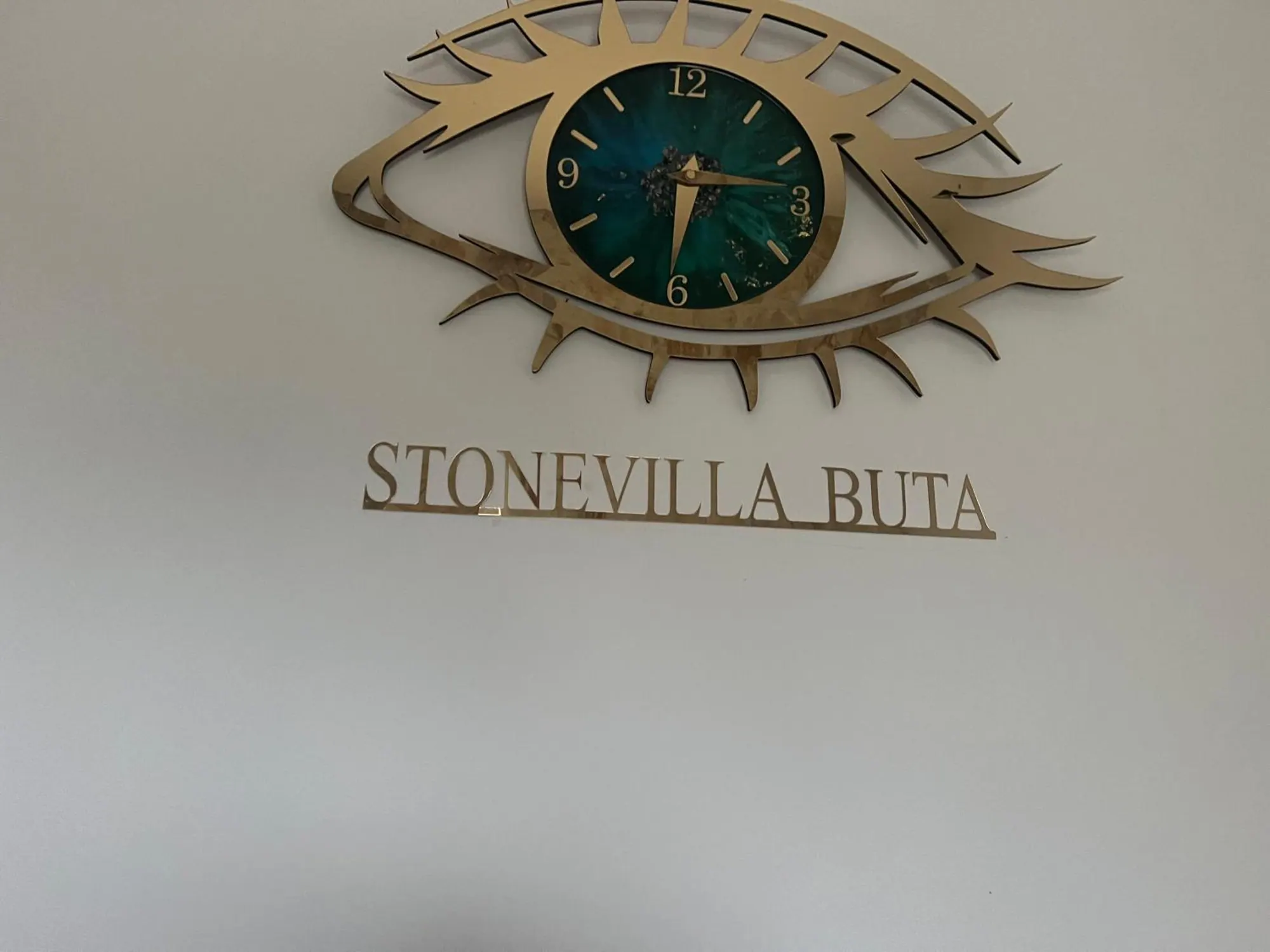 A brass clock in the shape of an eye, with a green resin face, above the name Stonevilla Buta in brass letters on a white wall