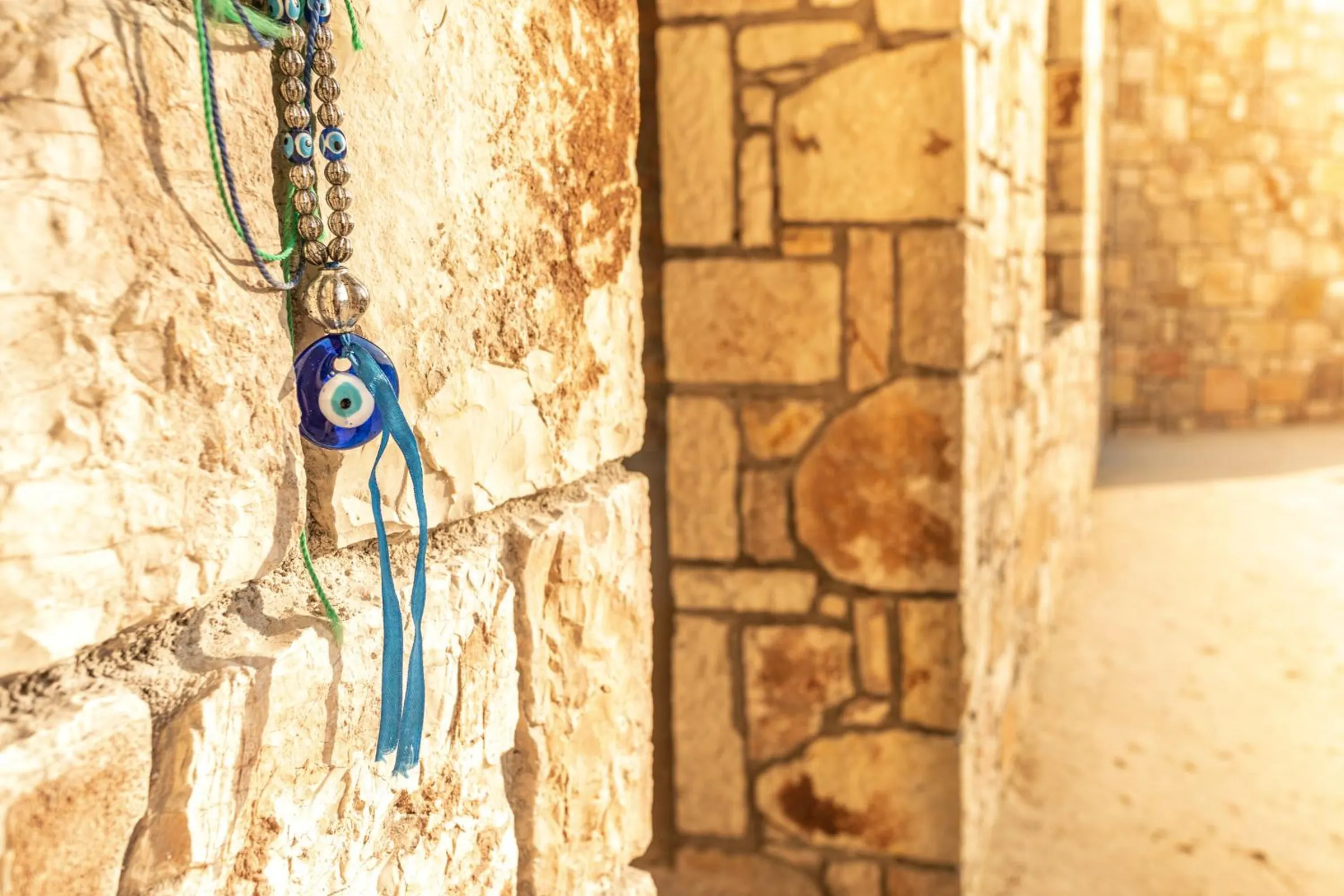 A blue glass eye charm on beads and ribbon, hung on the sunlit stone at the corner of the house