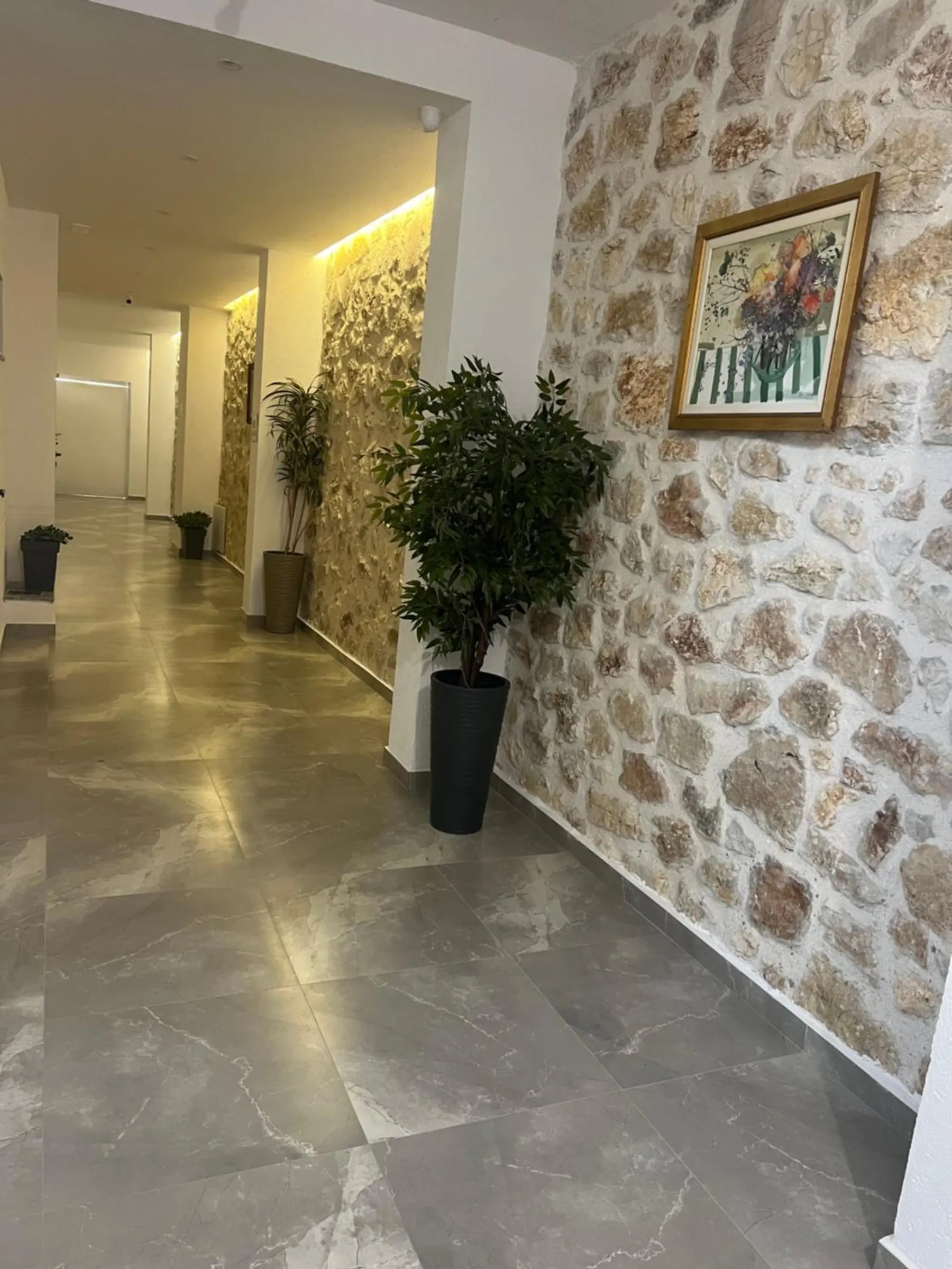 An upstairs hallway with grey marble floor, a stone wall hung with a painting, and a potted tree