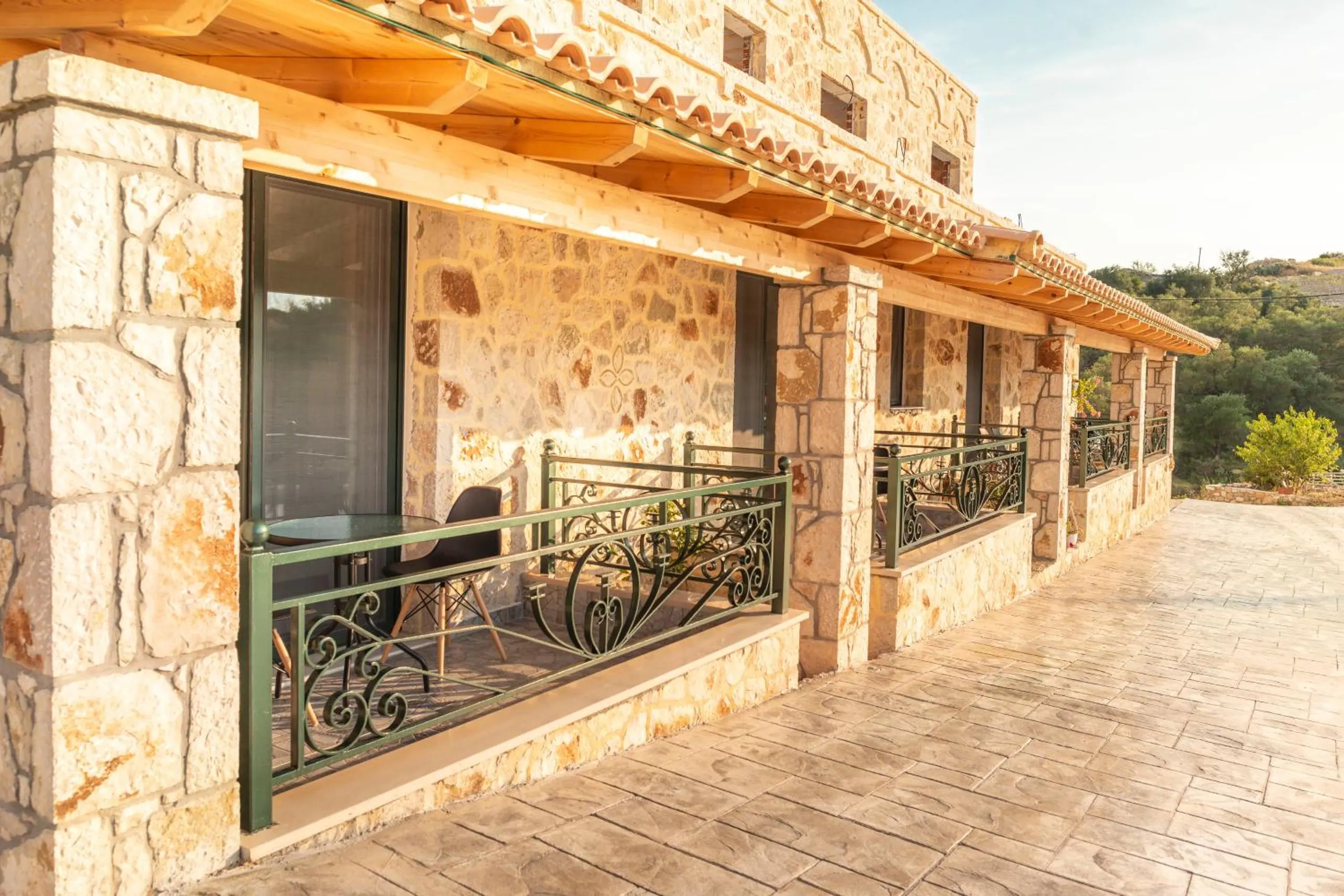 The ground-floor gallery in evening sun: stone pillars, a timber roof under terracotta tiles, and green wrought-iron balcony rails in front of each room