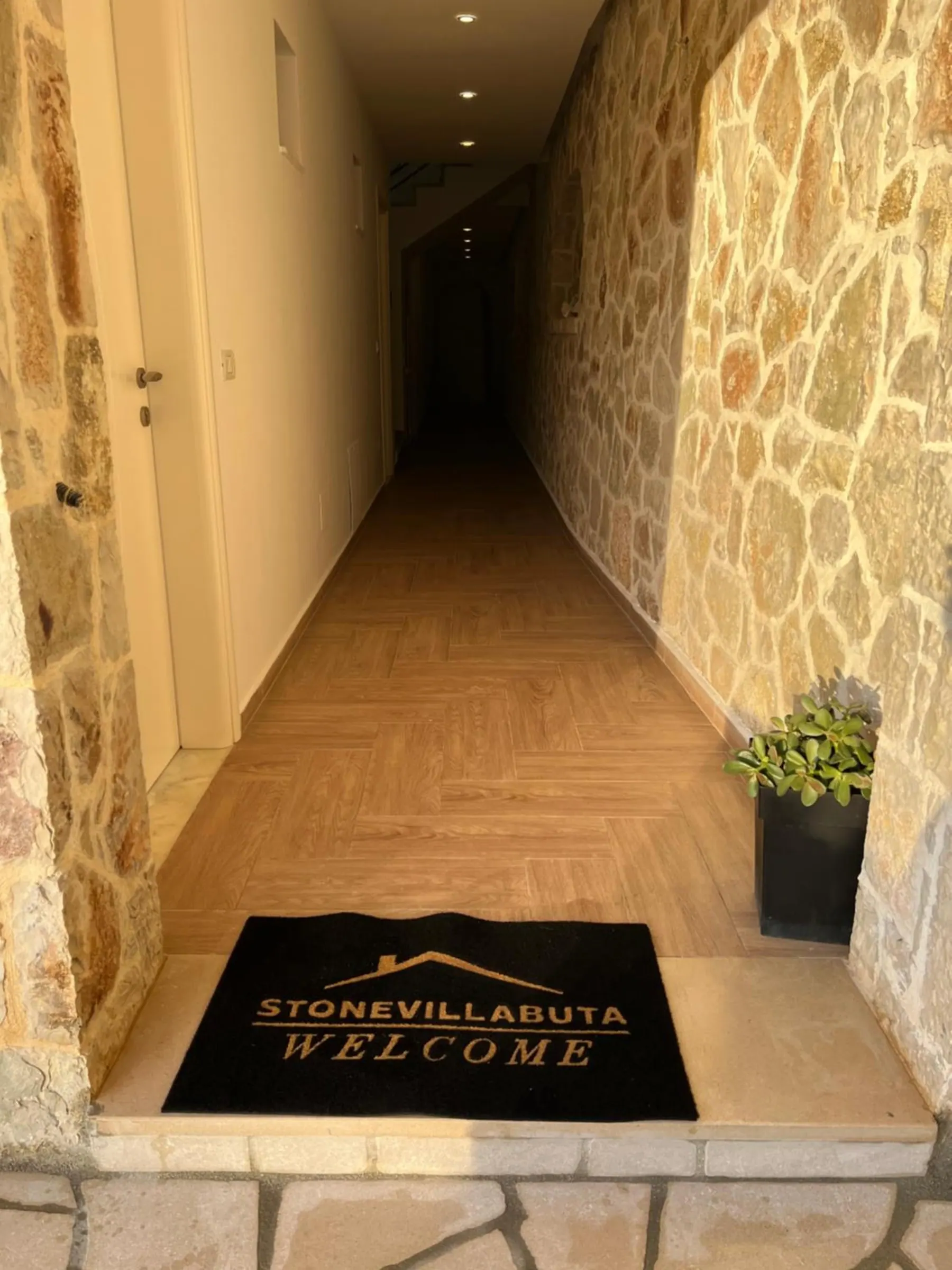 A corridor with a stone wall on one side, and at its door a black mat reading Stonevillabuta, Welcome