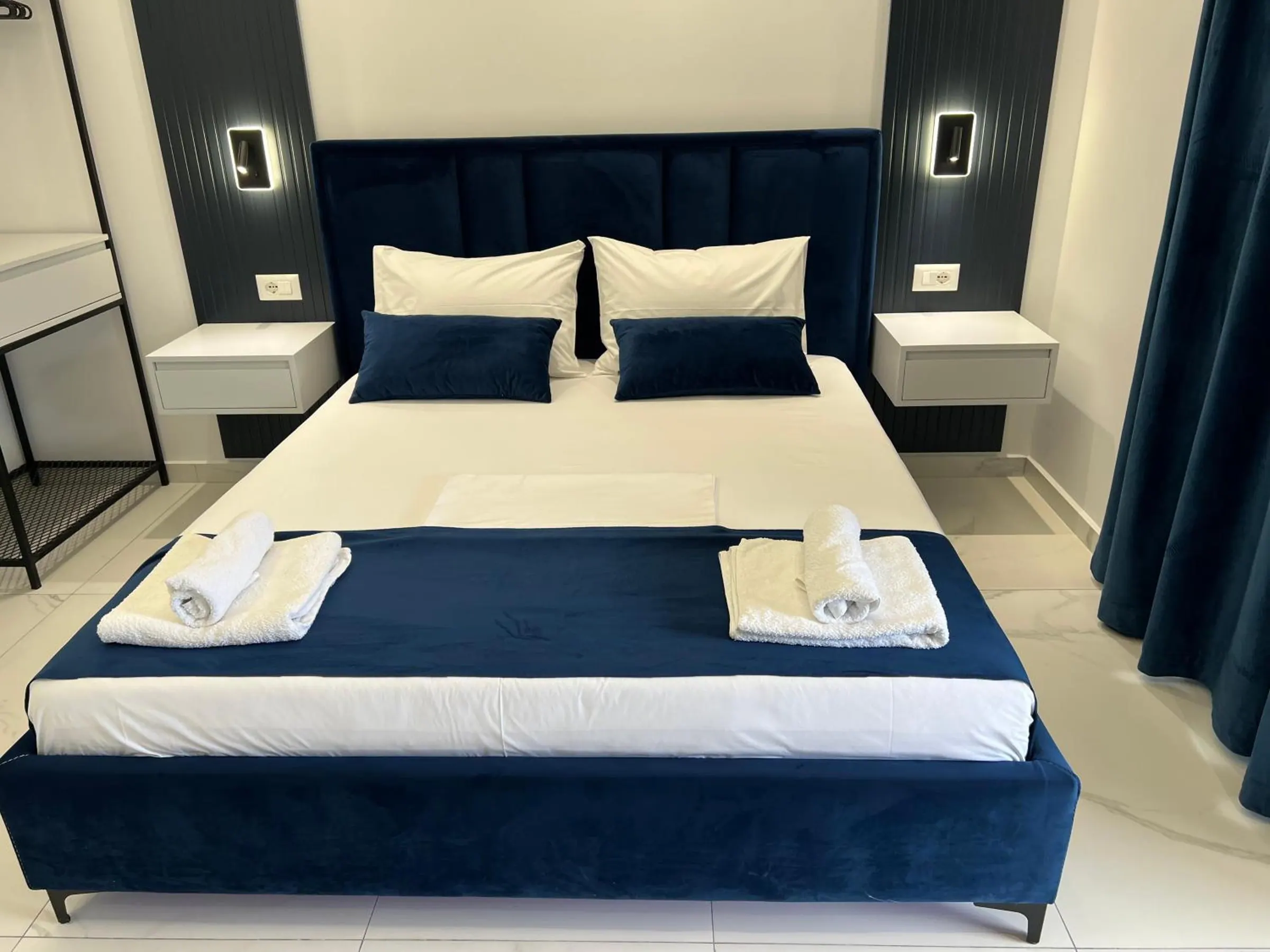 A double bed in navy velvet between two floating bedside tables, navy curtains to one side
