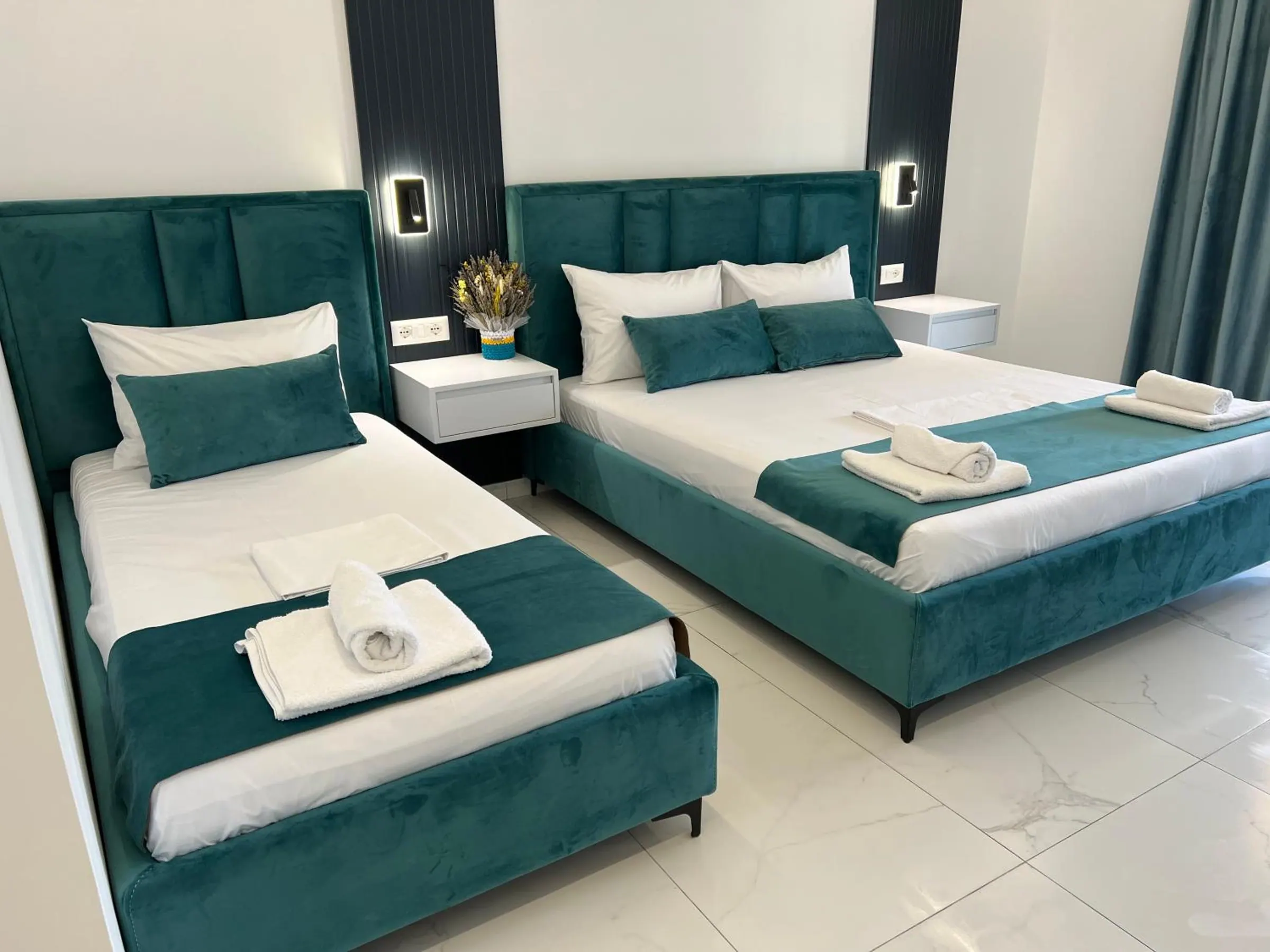 A single and a double bed in teal velvet with matching cushions, and white bedside tables