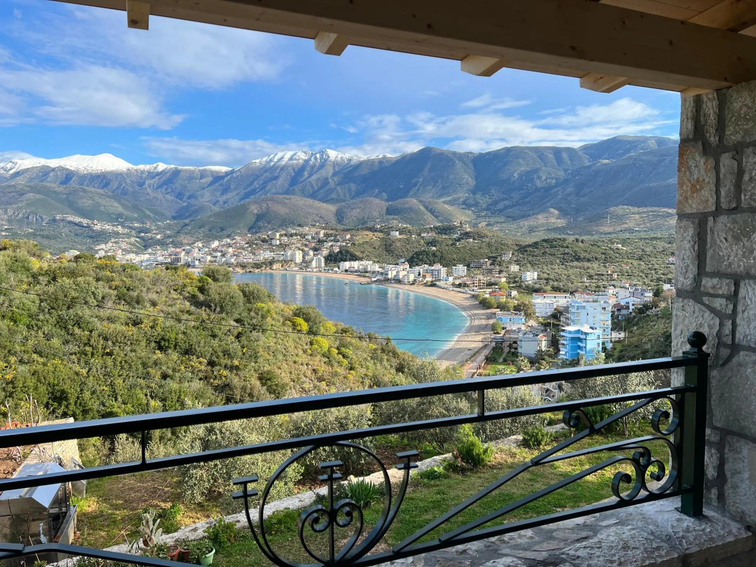 Potam bay from a balcony at Stonevilla Buta: a stone pillar and a wrought-iron rail in front, olive slopes falling to a turquoise curve of beach, the town beyond and snow on the mountains