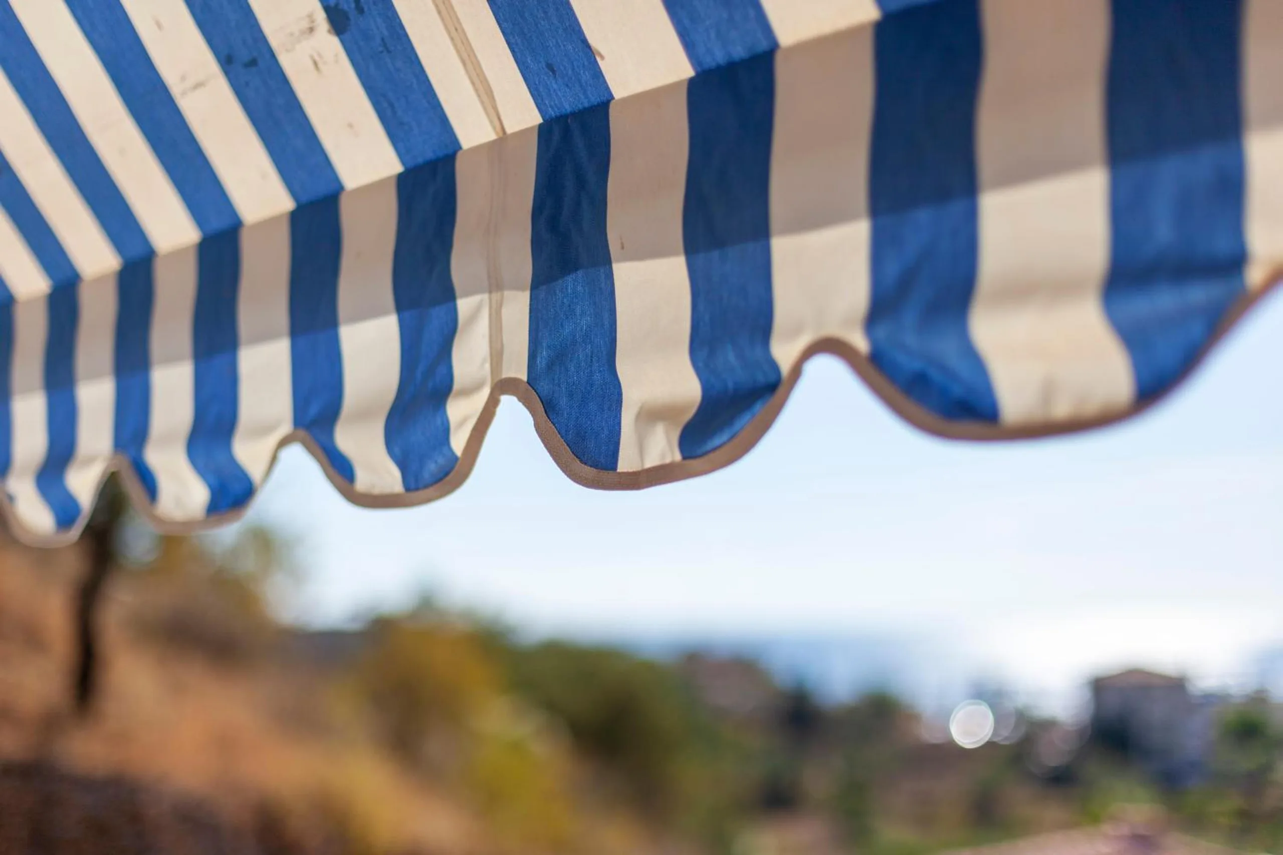 Close view of the blue-and-cream striped awning and its scalloped hem, with the hillside soft behind it