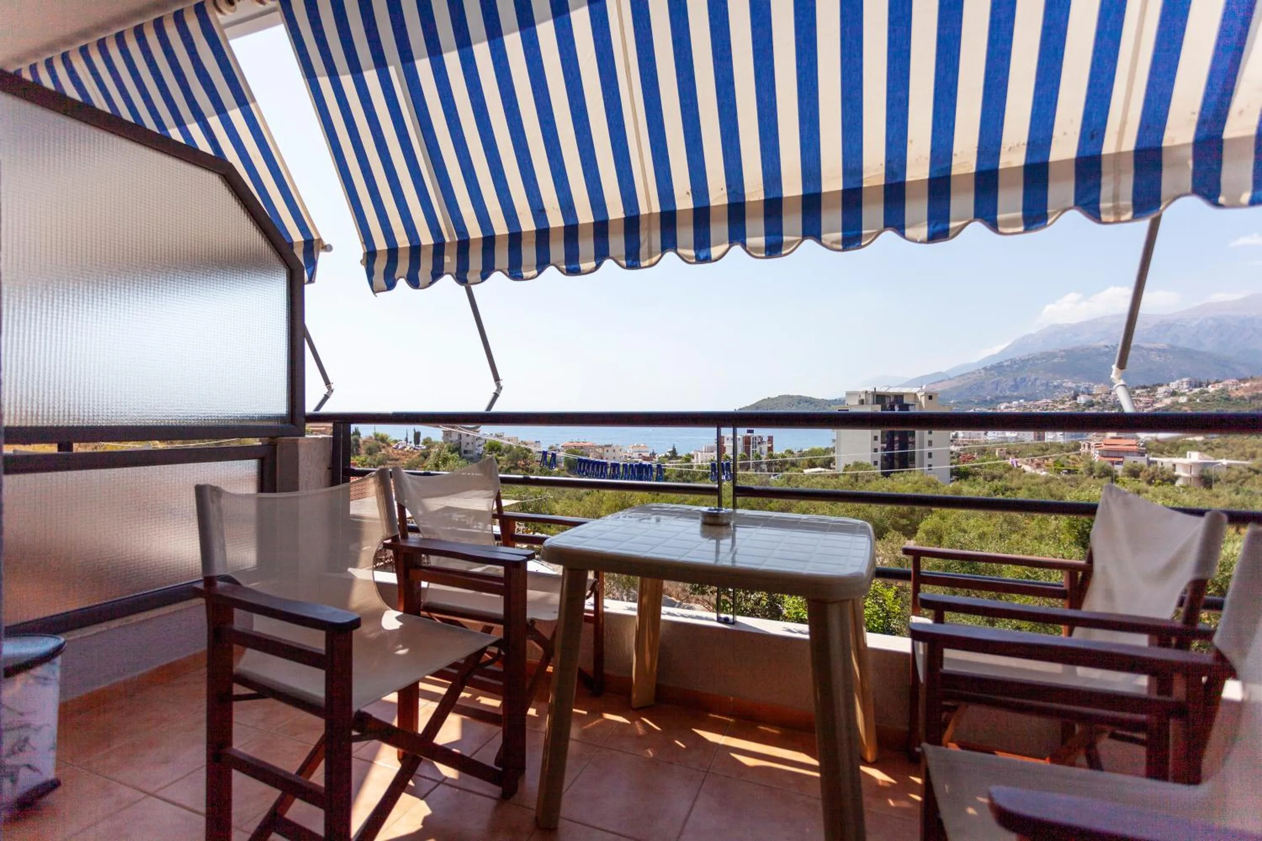 A balcony with a table and two director's chairs under a blue-and-cream striped awning, the olive groves, the town and the mountains beyond the rail