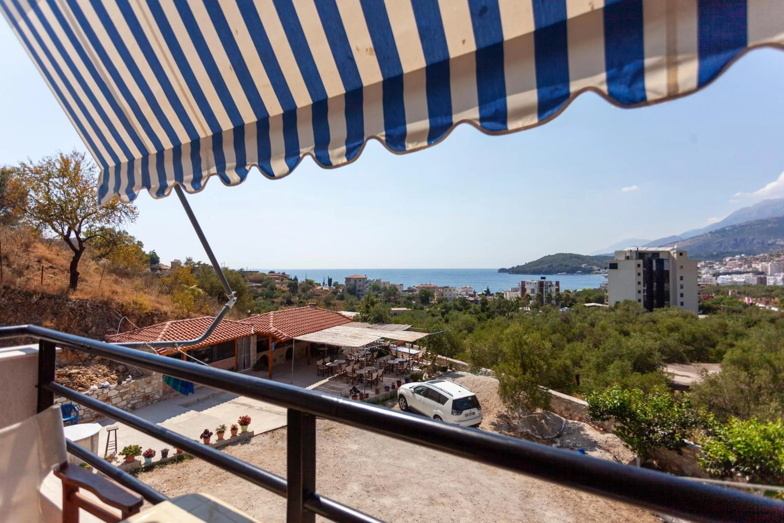 The scalloped edge of a blue-and-cream striped awning over a balcony rail, looking down over the terrace roof and the olive trees to the bay and the headland