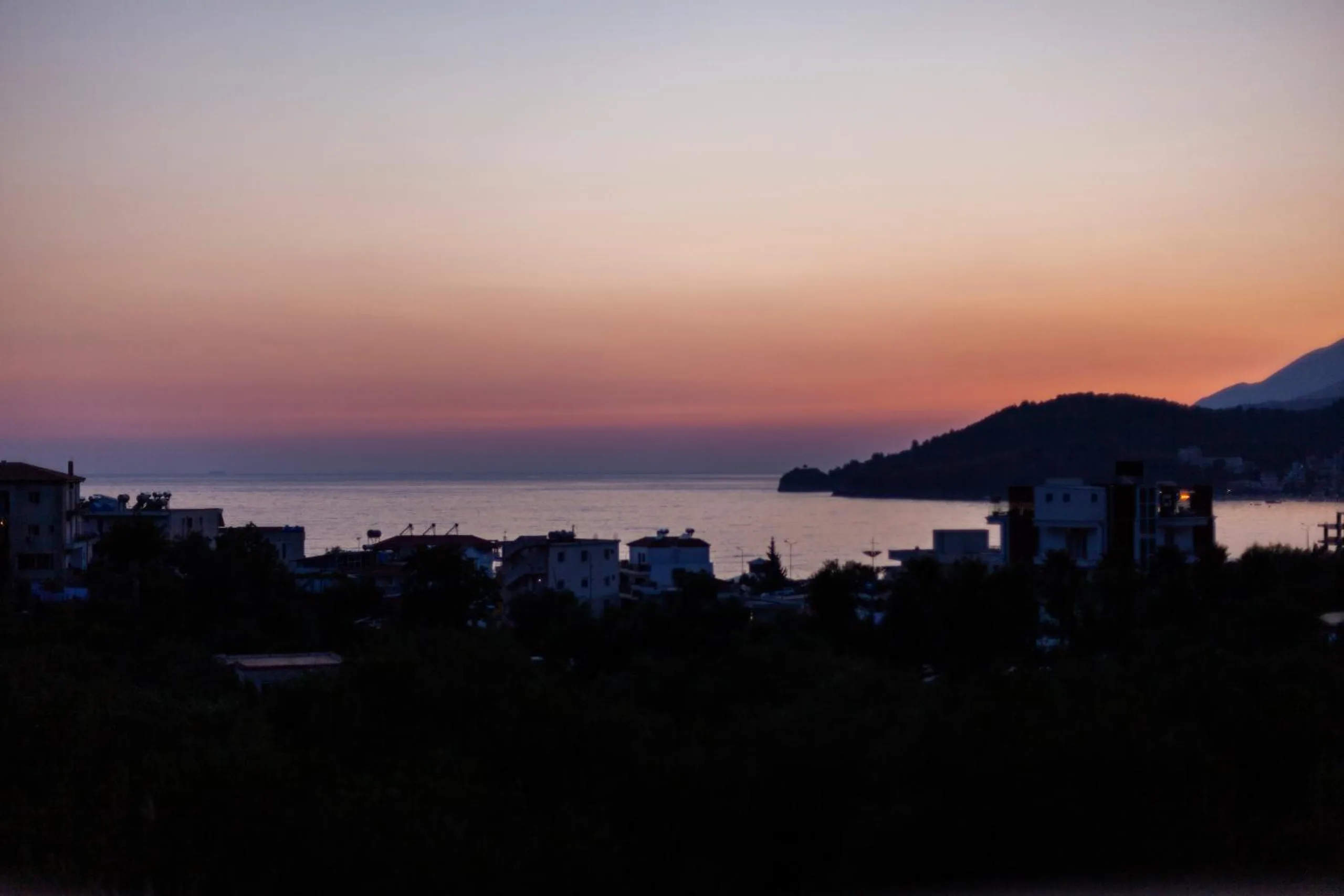 Himarë Bay after sunset, the headland dark against an orange and violet sky and the sea still pale