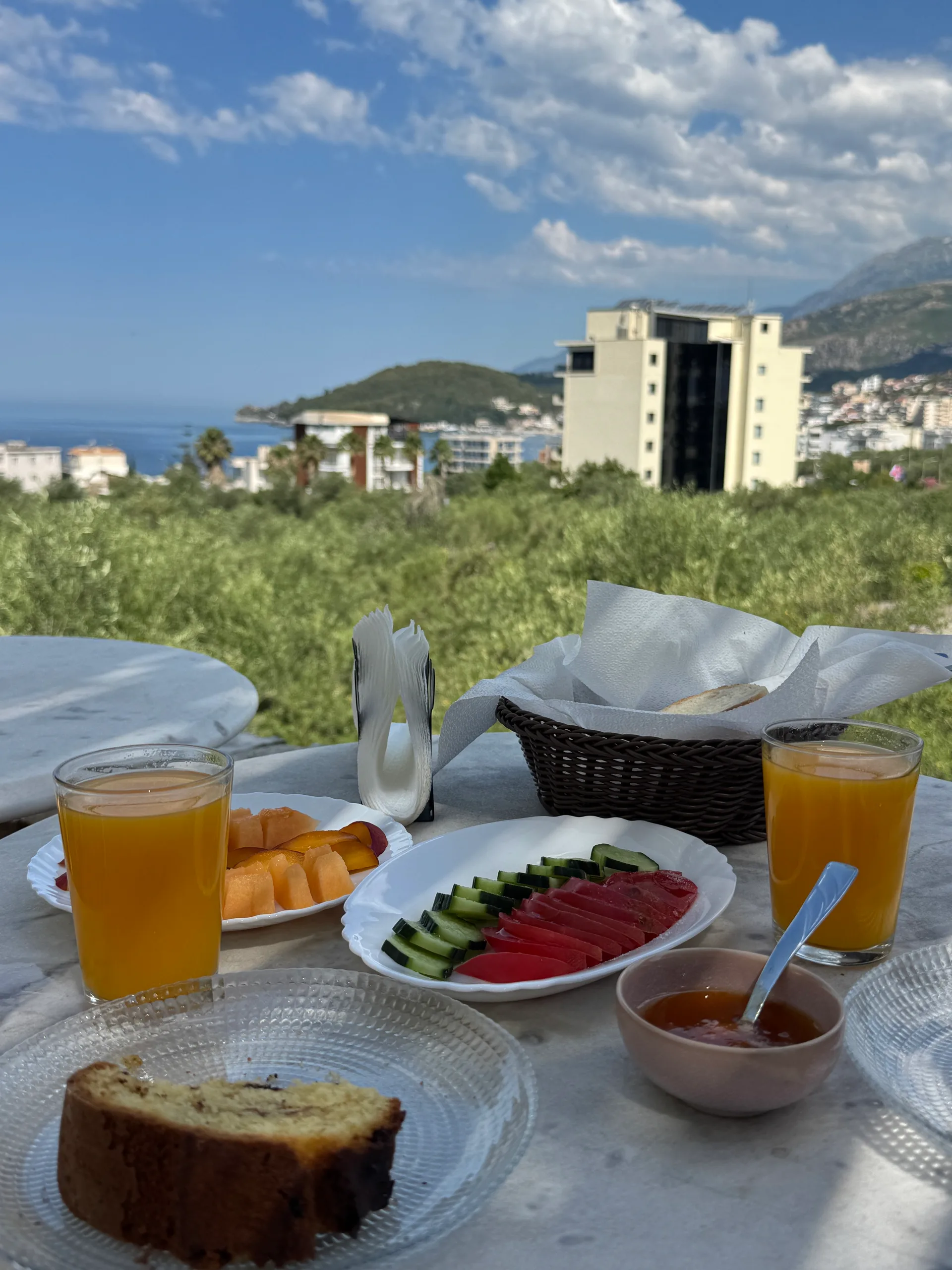 Breakfast on a marble table: two glasses of orange juice, sliced melon, tomato and cucumber, a bread basket, a slice of cake and a small bowl of honey, with the olive groves and Himarë Bay behind