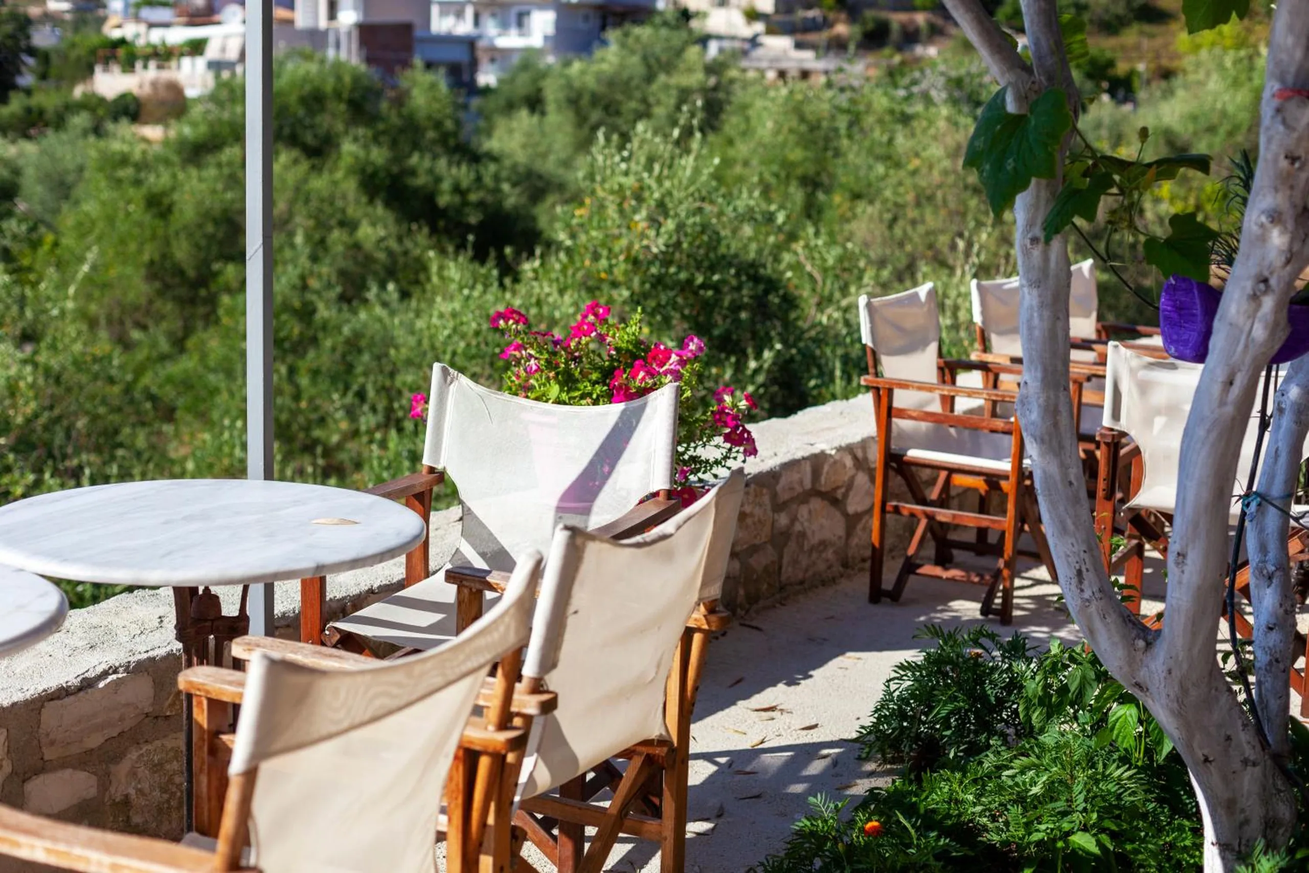 A marble table and canvas chairs on a stone-walled garden terrace, pink geraniums and olive branches overhead and houses on the hill below