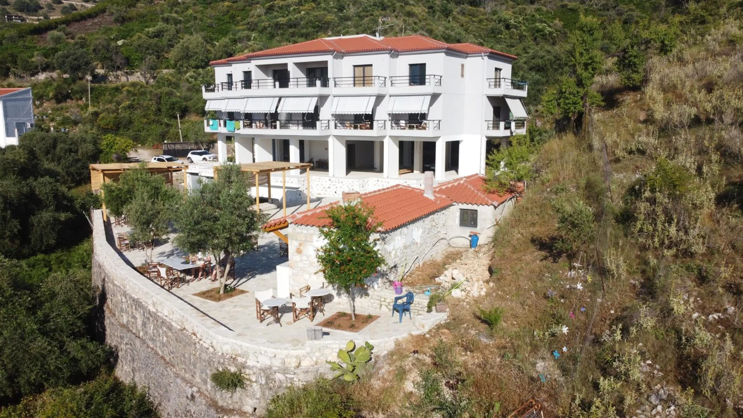 The house from the air: a white three-storey building with a terracotta roof and striped awnings over the balconies, a lower stone cottage, and a terrace of tables and olive trees held up by a curved dry-stone wall on the hillside