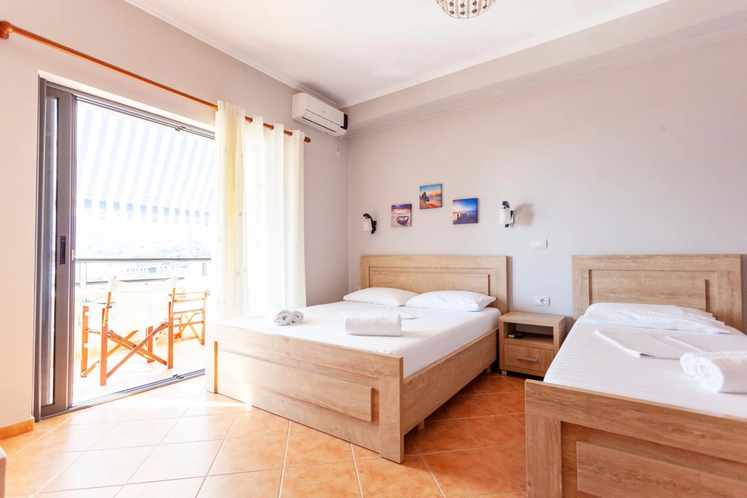 A bright room with a double and a single bed in pale oak, terracotta floor tiles and a glass door open onto a balcony under a striped awning