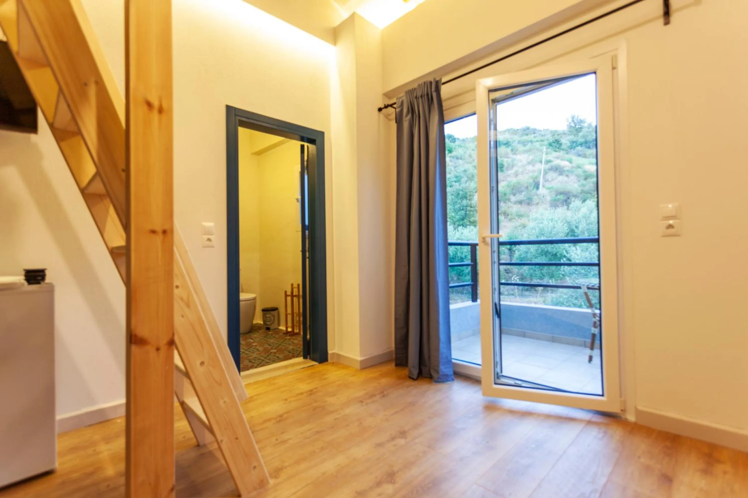 A pine ladder climbing to a sleeping loft, a blue-framed bathroom door and a glass door open onto a balcony over the olive trees