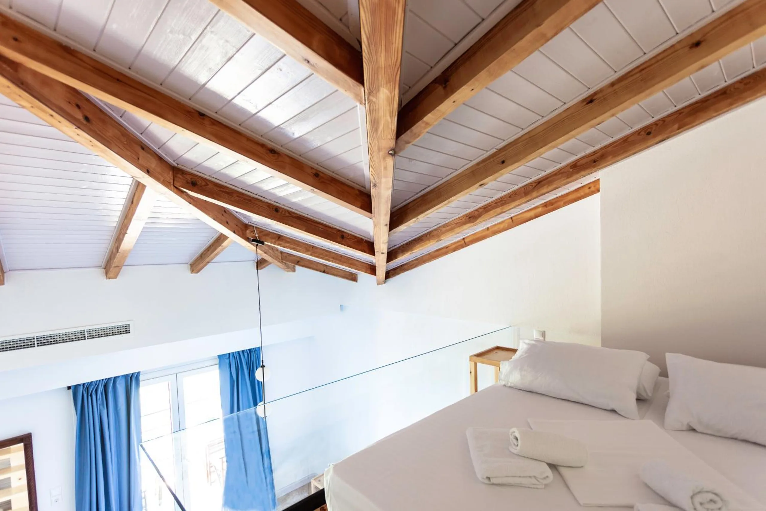 A bed on a mezzanine under white boarded ceiling and exposed timber trusses, a glass balustrade at its edge and blue curtains at the window below