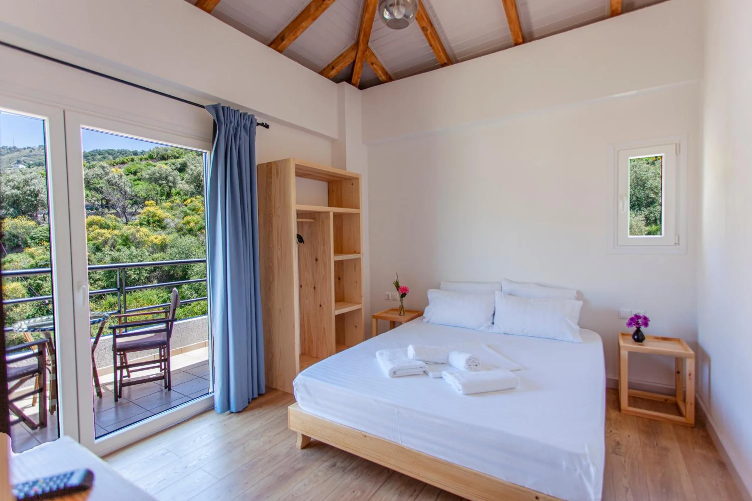 A room under the roof with a pine bed, open shelving and blue curtains, glass doors open onto a balcony with director's chairs and the green hillside