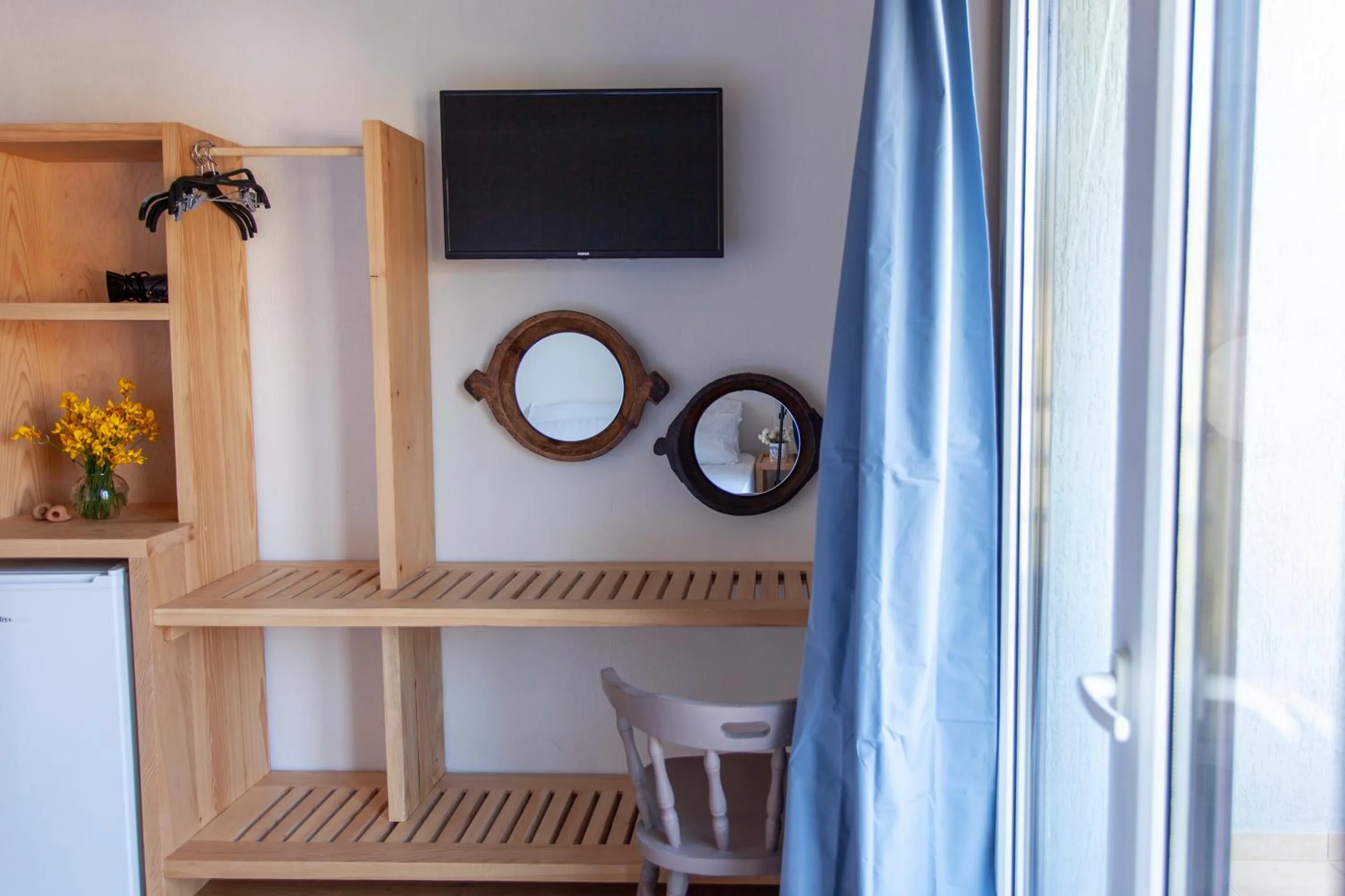 Pine shelving with a small fridge and a television, two round wooden porthole mirrors on the wall, a vase of yellow flowers and a blue curtain at the window