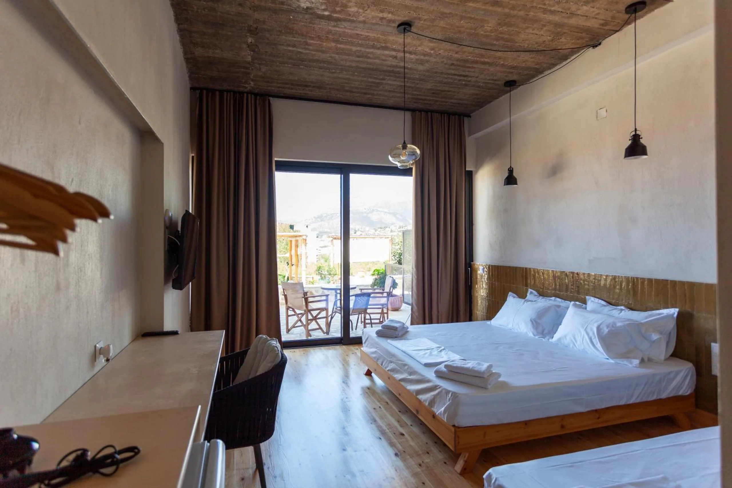 A long room with a dark timber plank ceiling, a double bed on a low wooden frame, pendant lamps and full-height glass doors onto a terrace