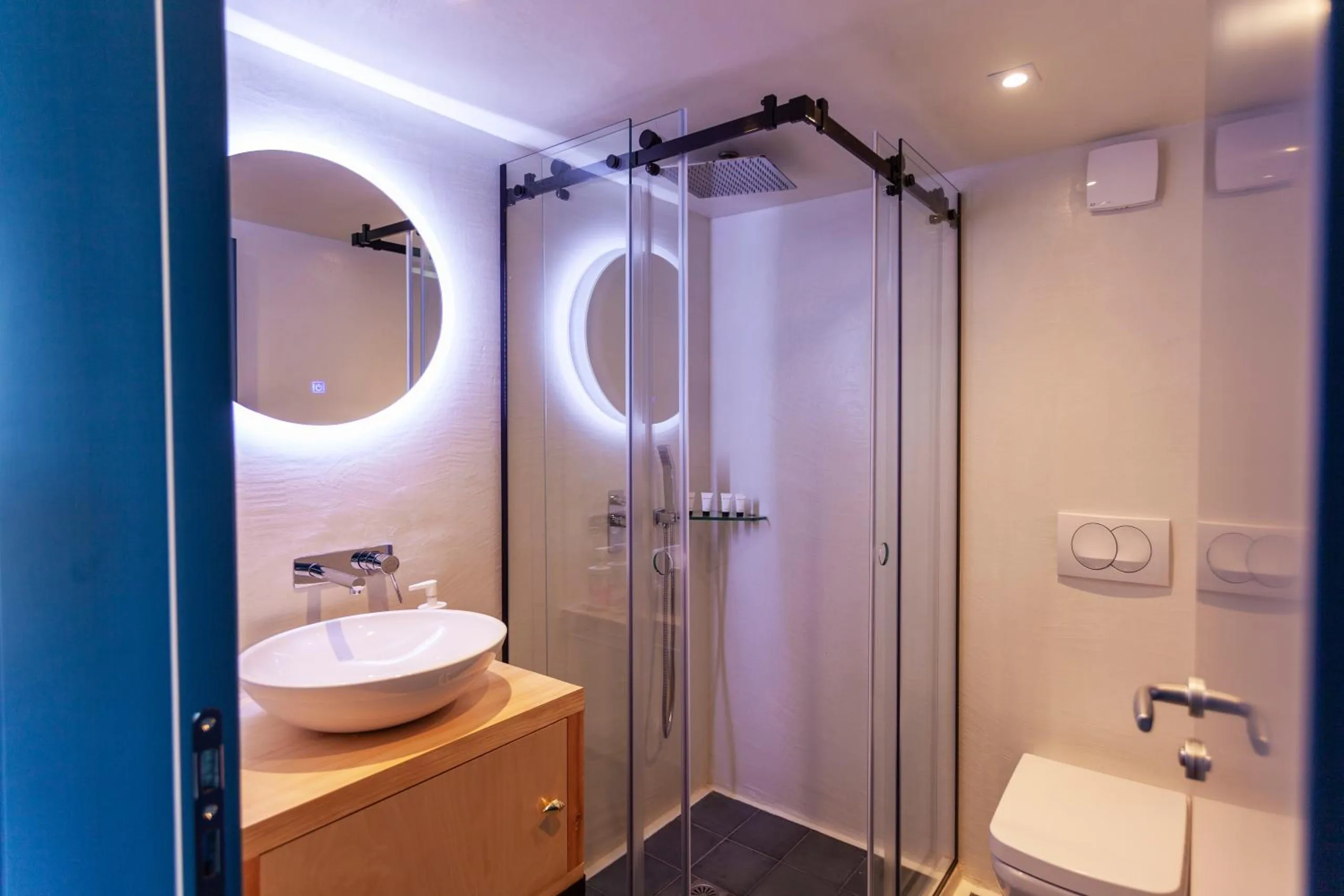 A bathroom with a round backlit mirror over a white bowl basin on a pine stand, a glass shower and a blue door frame