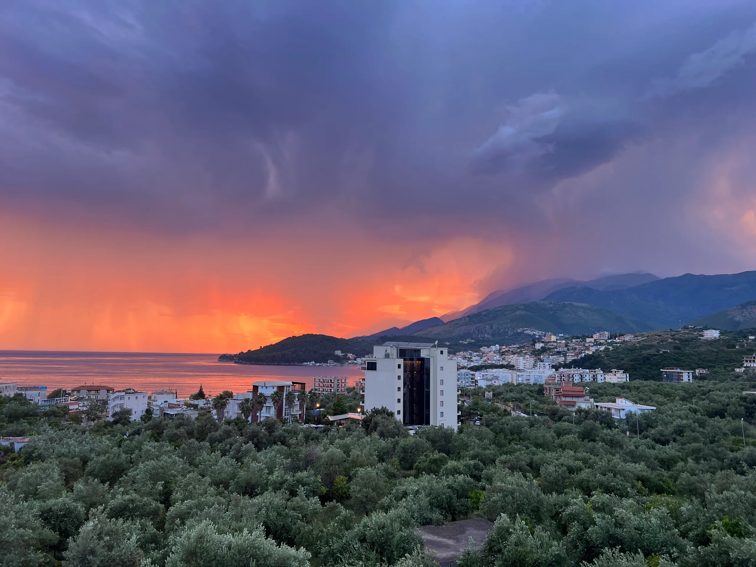 A storm passing over Himarë Bay at sunset, orange light under violet cloud, the olive groves dark in the foreground and the town along the shore
