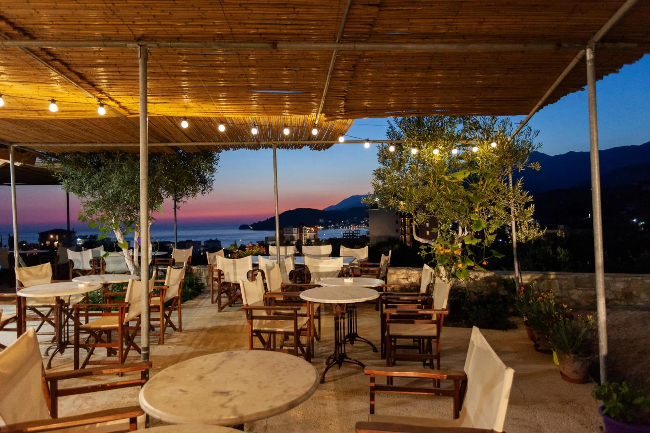 The terrace at dusk under a reed roof strung with bulbs: marble bistro tables and canvas director's chairs among olive trees, the bay and its headland below against a pink and violet sky