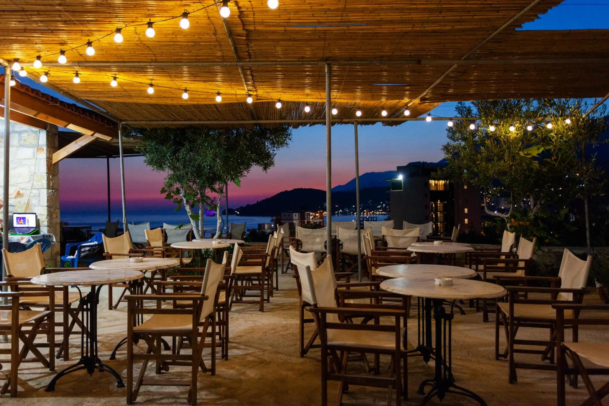 Rows of canvas director's chairs and round marble tables under a reed pergola hung with lit bulbs, a stone wall on the left and the lights of Himarë across the bay
