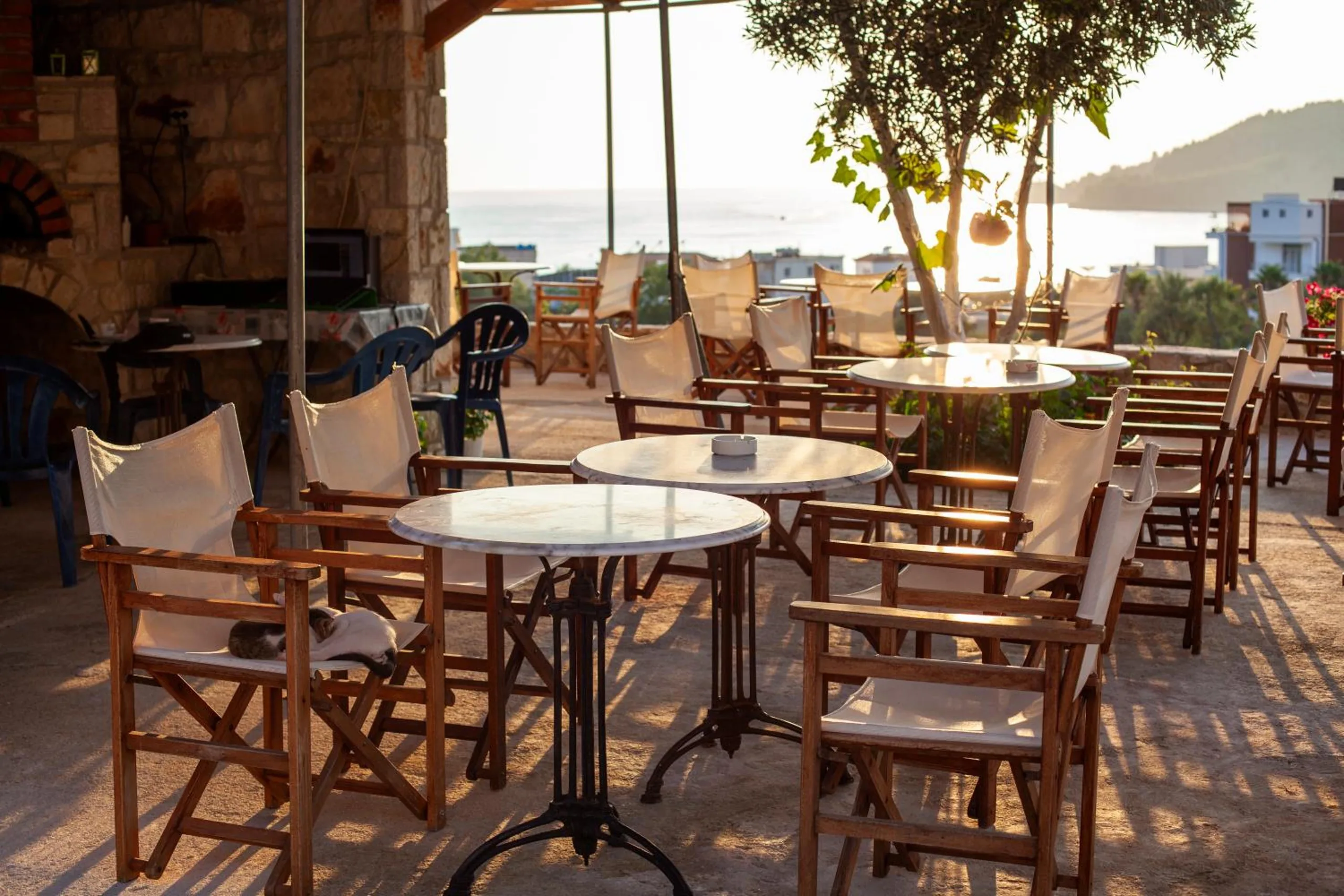 The corner of the terrace by a stone-built wall with an arched oven, marble tables and wooden chairs in evening light and the sea beyond