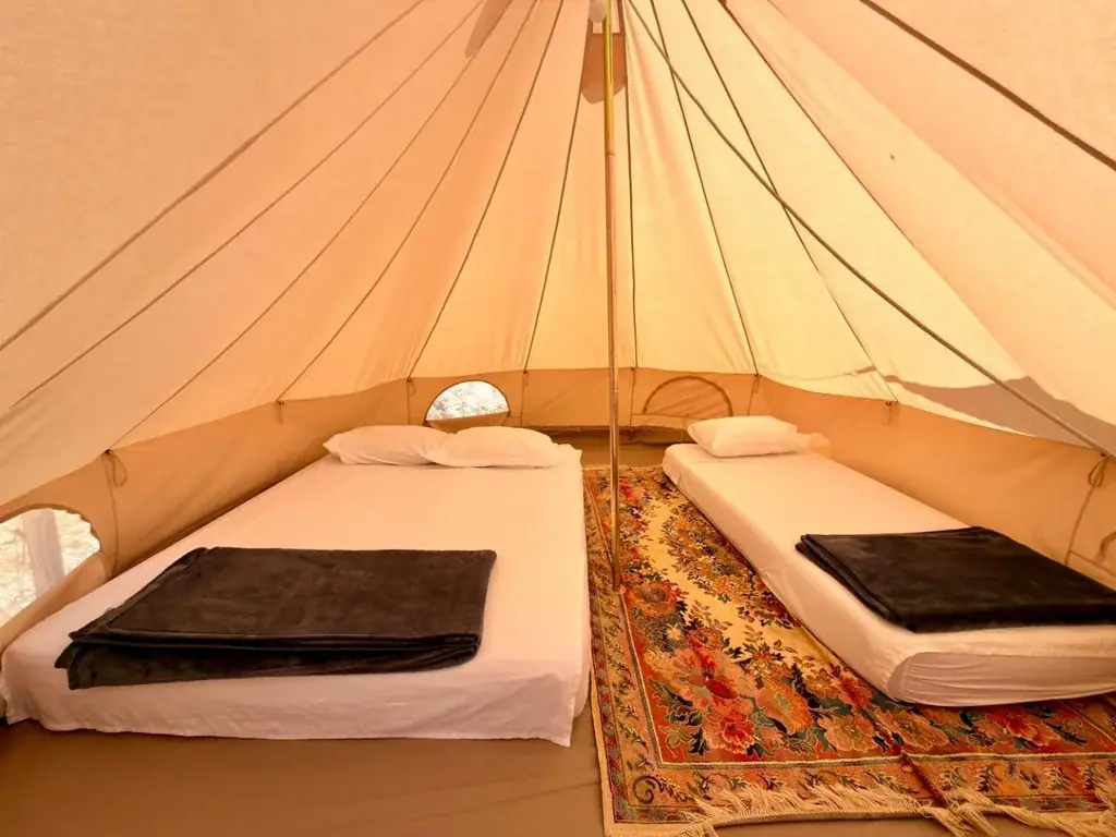 Inside a bell tent at Albania Glamping: a double bed and a single bed in white linen with folded brown blankets, a floral rug between them, and the timber centre pole under the canvas