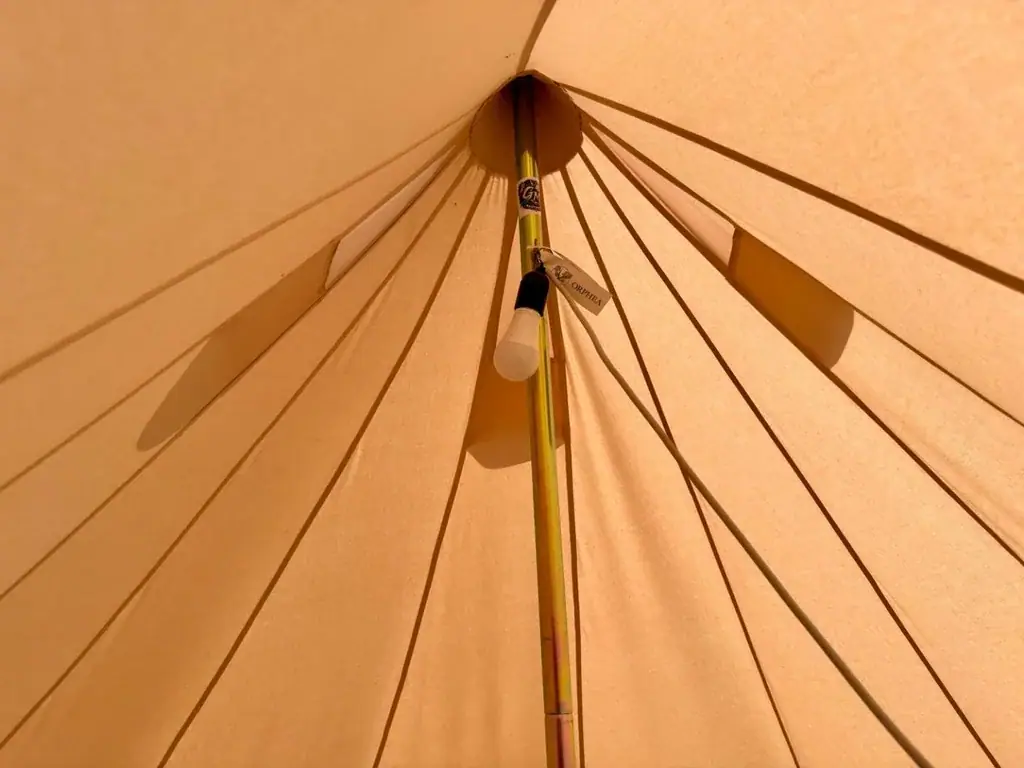 Looking up at the crown of a bell tent at Albania Glamping: canvas gores radiating from the peak around a brass-toned centre pole with a single bare bulb hanging from it