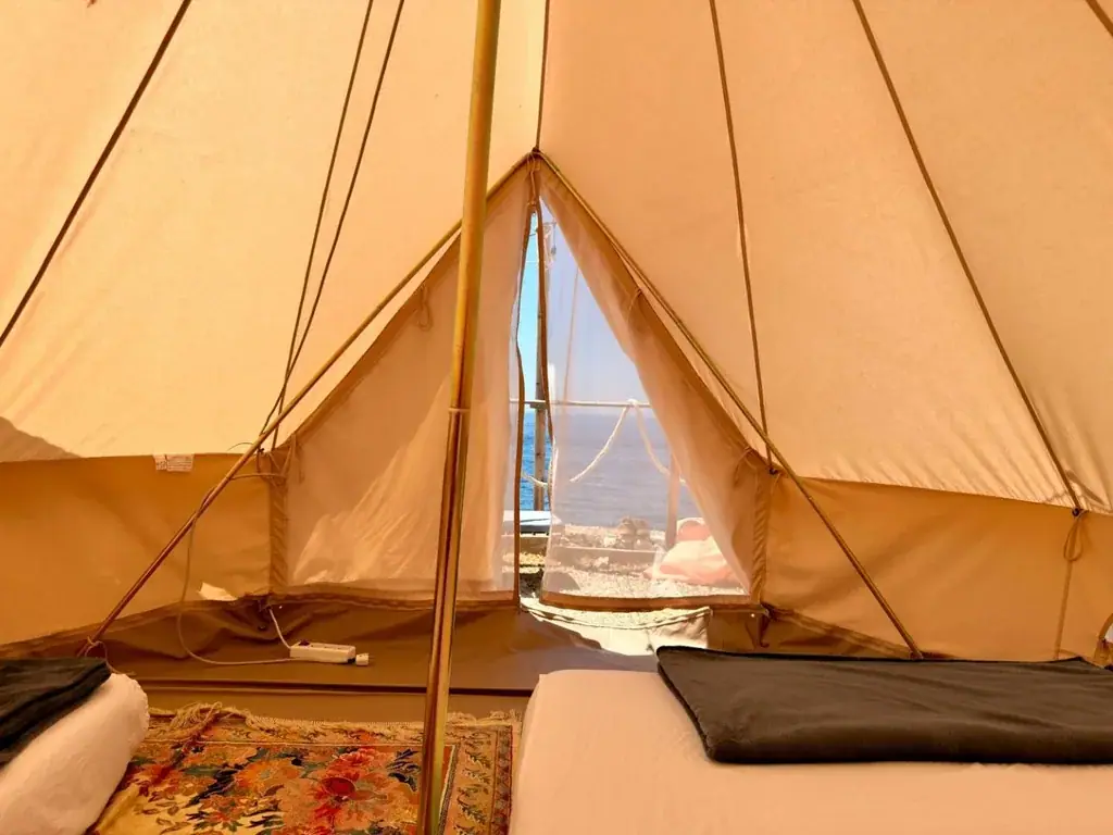 The open doorway of a bell tent at Albania Glamping with the mesh screen down, looking out past a rope railing and an orange bean bag to the flat open sea