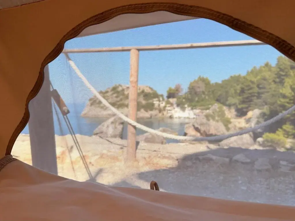 The crescent mesh window of a tent at Albania Glamping framing the cove outside: pale rock, a rope railing on timber posts and a pine-covered headland across the water