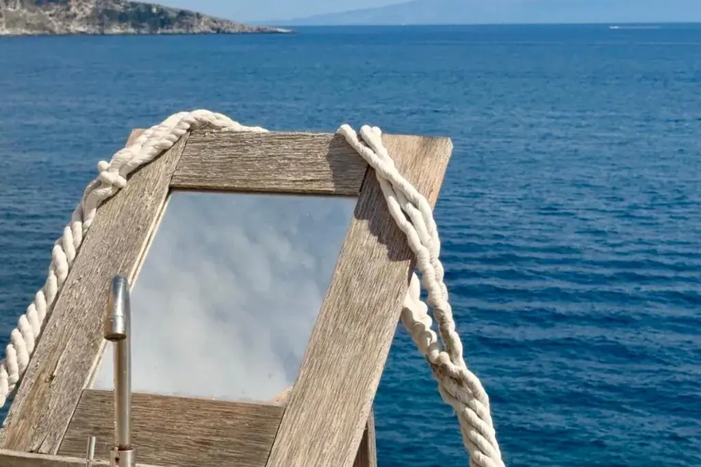 A driftwood-framed mirror lashed with white rope to a post at Albania Glamping, a chrome tap beneath it, with the open Ionian and a rocky headland behind