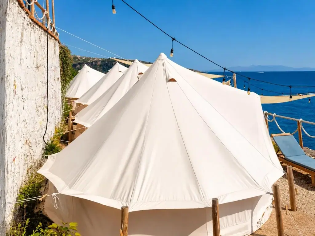 Four white bell tents guyed to timber posts along a whitewashed retaining wall at Albania Glamping, under a run of festoon bulbs and a shade sail, with a sun lounger and the open sea beyond
