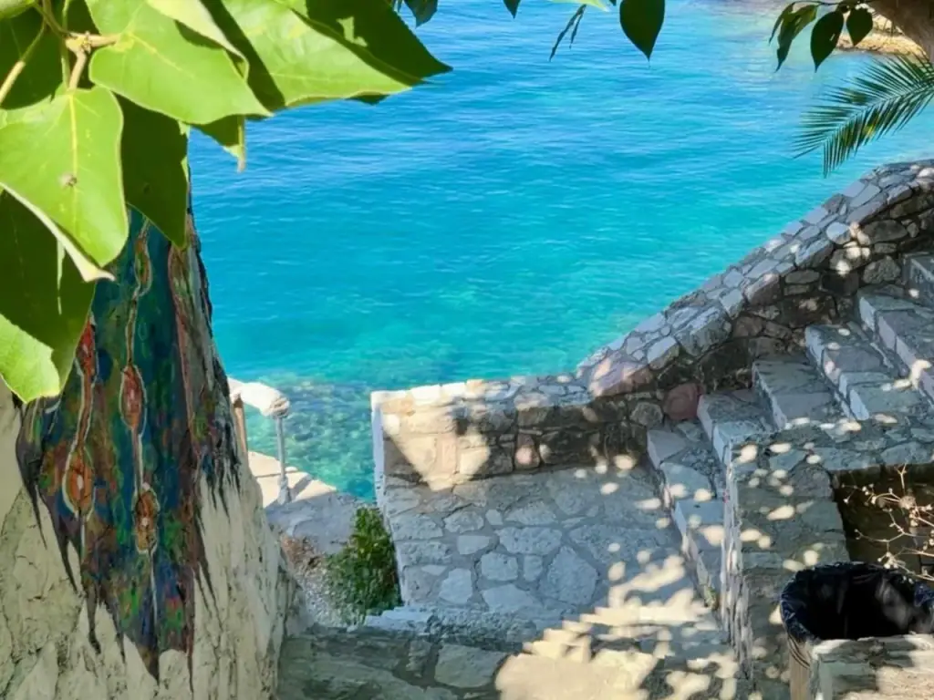 Whitewashed stone steps at Albania Glamping dropping between dry-stone walls to clear turquoise shallows, with a painted panel on the wall and fig leaves overhead