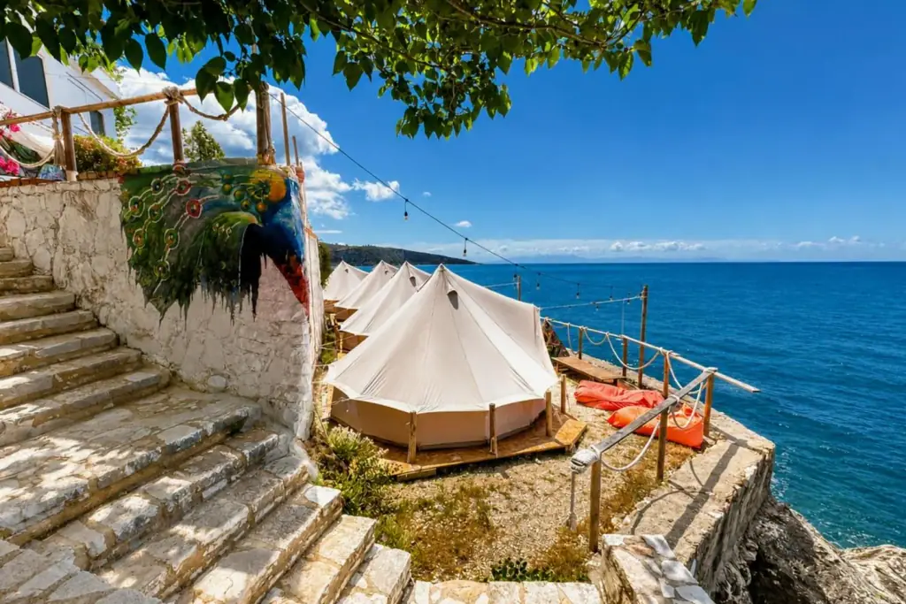 A row of white canvas bell tents on timber decks along a narrow stone terrace above the Ionian at Albania Glamping, with a painted peacock mural on the whitewashed wall and festoon lights strung over the rope railing