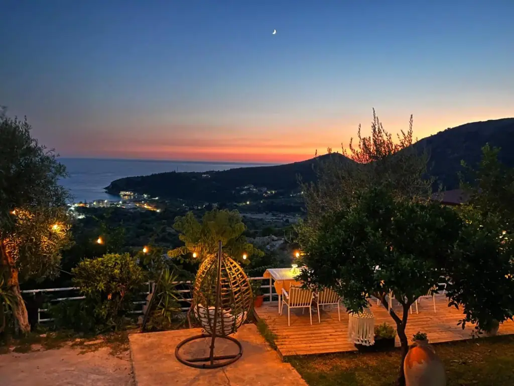 A timber deck at dusk with a hanging wicker chair and lit trees, the lights of the bay far below and an orange horizon