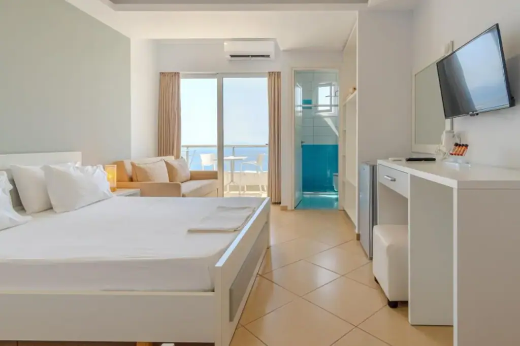 A white room with a double bed and a sliding glazed door onto a balcony, the sea in the opening