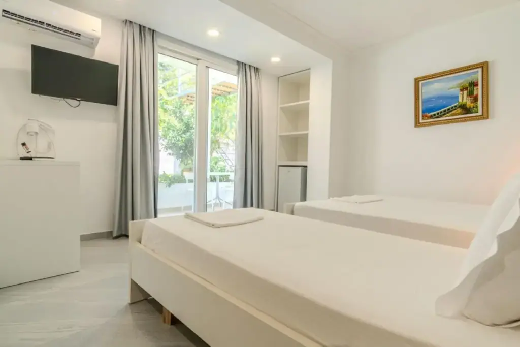 A room with a bed, a wall-mounted television and a framed picture, the door open onto greenery