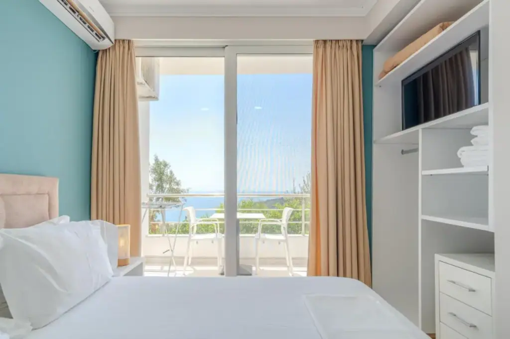 A room with a teal accent wall and cream curtains, a glazed door onto a balcony with two chairs and the sea