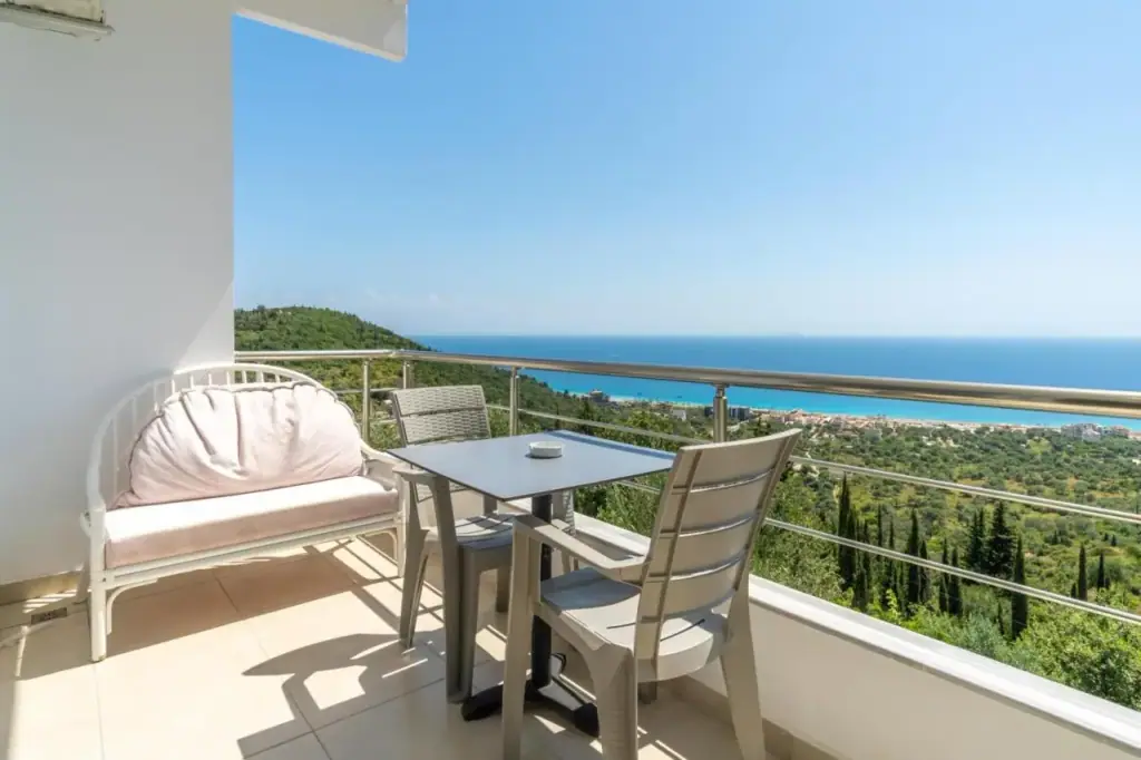 A white sofa, table and chairs on a balcony behind a steel rail, the whole bay and the sea beyond it