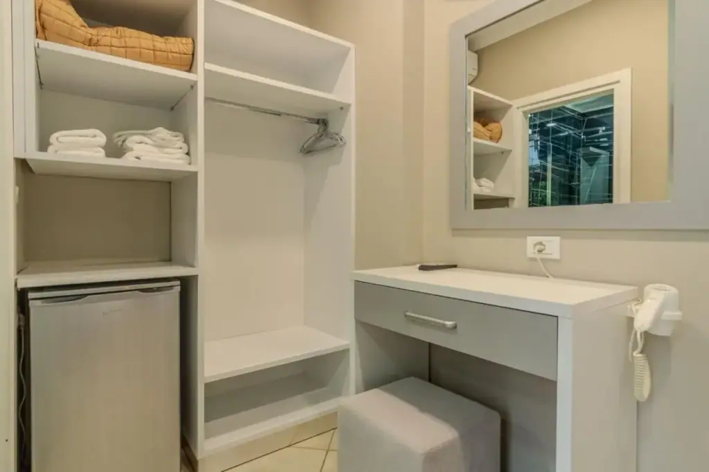 An open wardrobe with folded towels beside a white desk, a mirror, a hairdryer and a small refrigerator