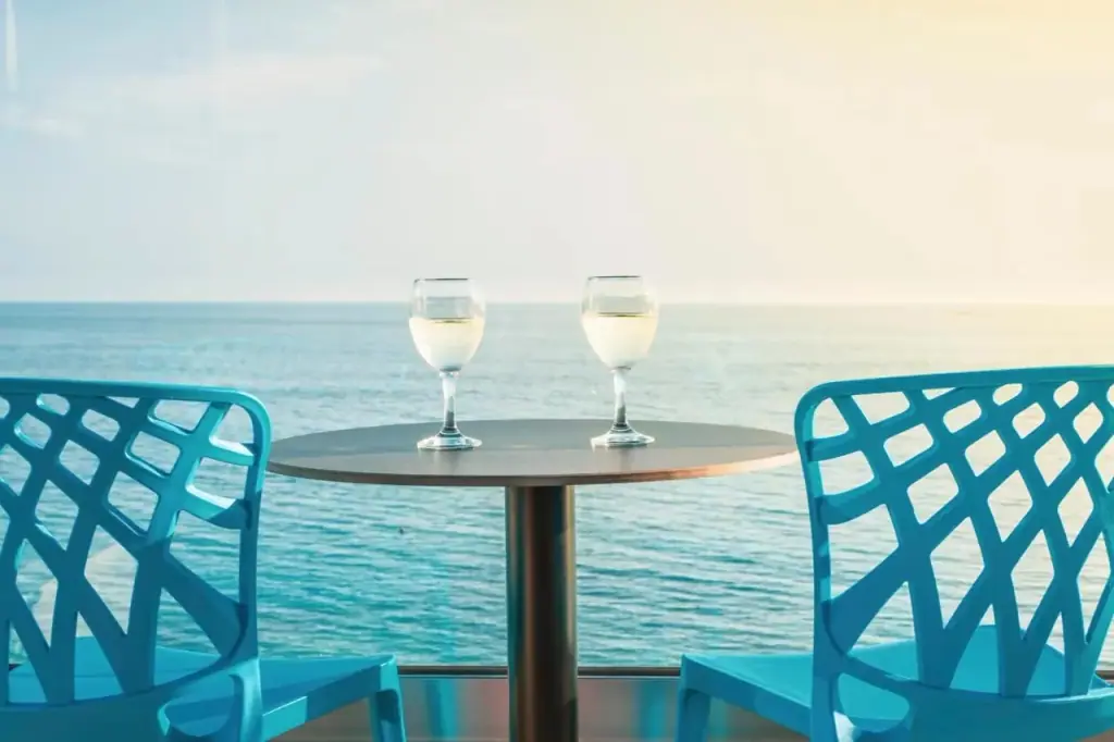 Two glasses of white wine on a balcony table between turquoise chairs, with the sea behind