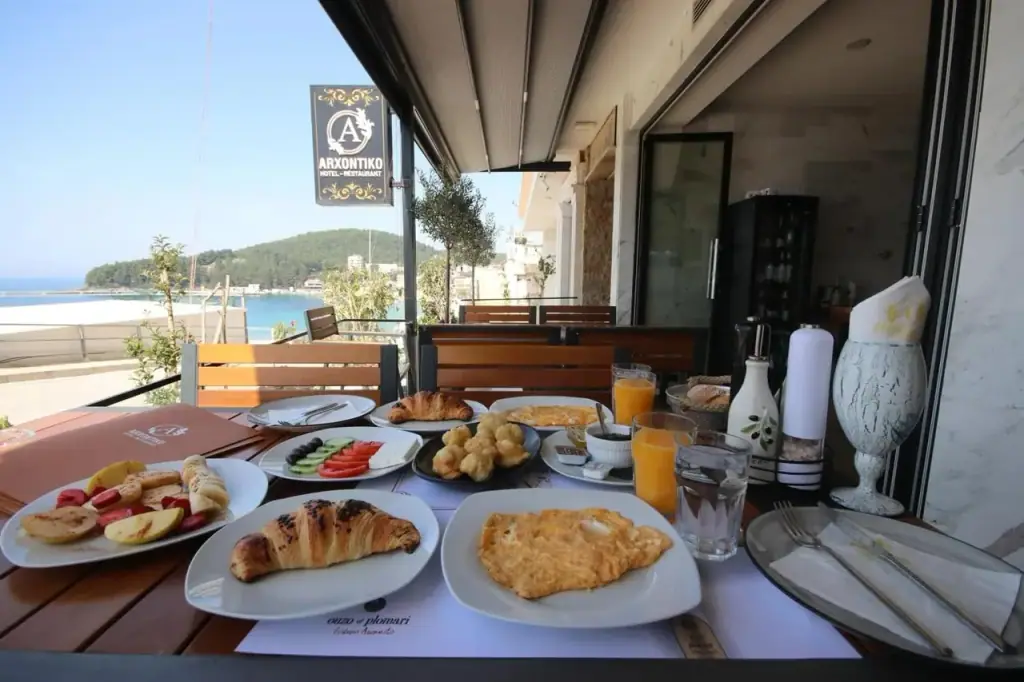 A breakfast table on the terrace with the black-and-gold ARXONTIKO plaque above it and Himarë bay behind