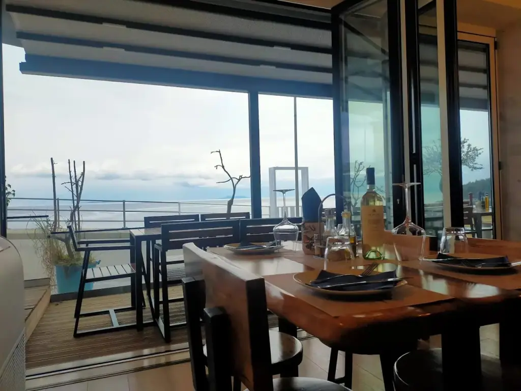 A laid table inside the restaurant, black plates and a bottle of white wine, with the deck and the open sea beyond the folded-back frontage