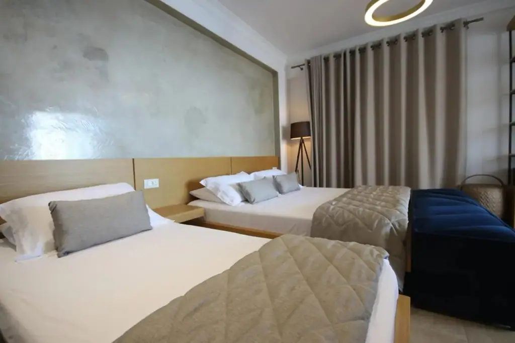Two beds side by side under quilted throws, with timber headboards under a panel of polished plaster and a standing lamp