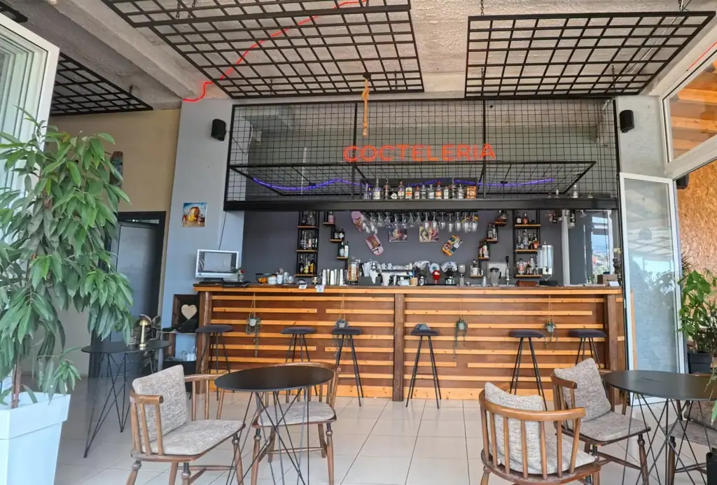 The bar counter in timber slats under a black steel mesh ceiling, with a red neon Cocteleria sign, stools along the counter and tables on the white tile floor