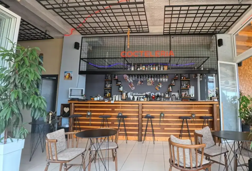 The bar counter in timber slats under a black steel mesh ceiling, with a red neon Cocteleria sign and a violet neon tube